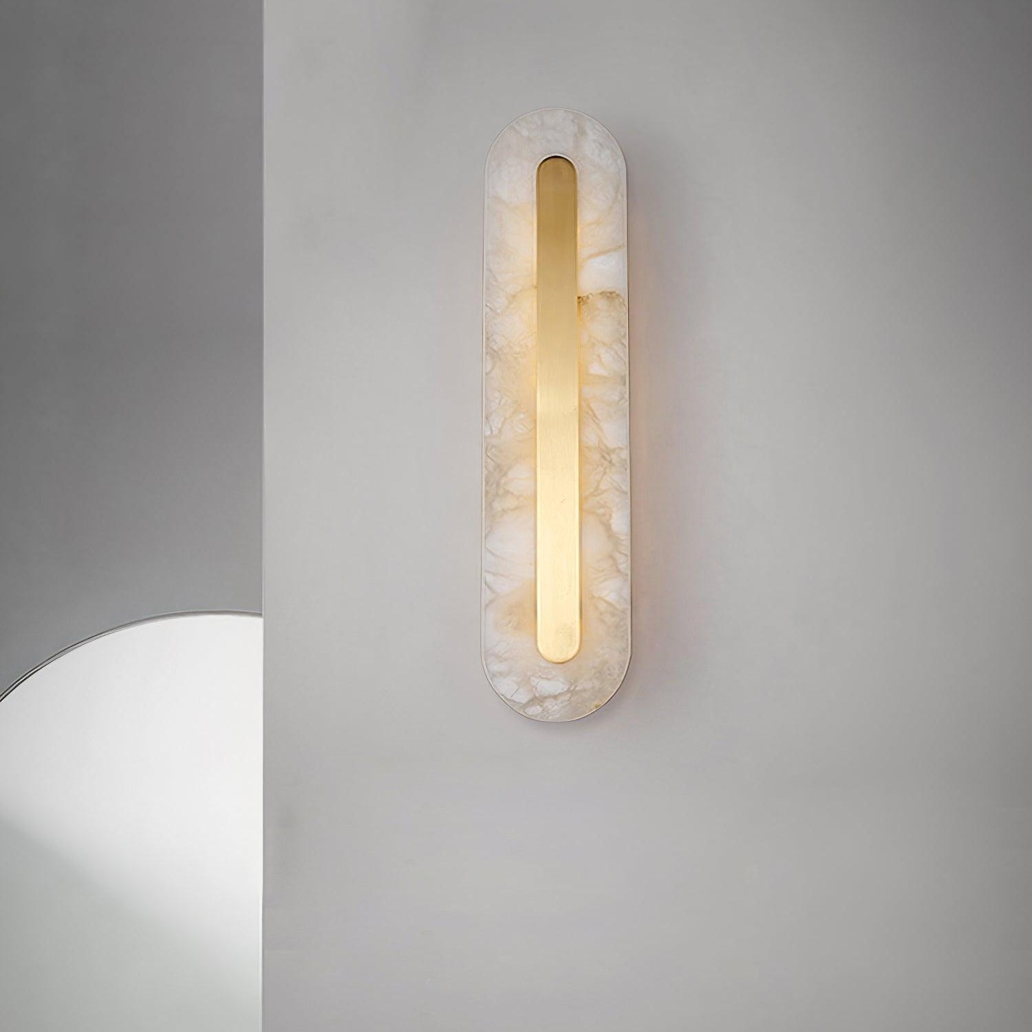 Clérachic selvia wall light lit on a pale grey corner wall, the curved edge of a round mirror at the left