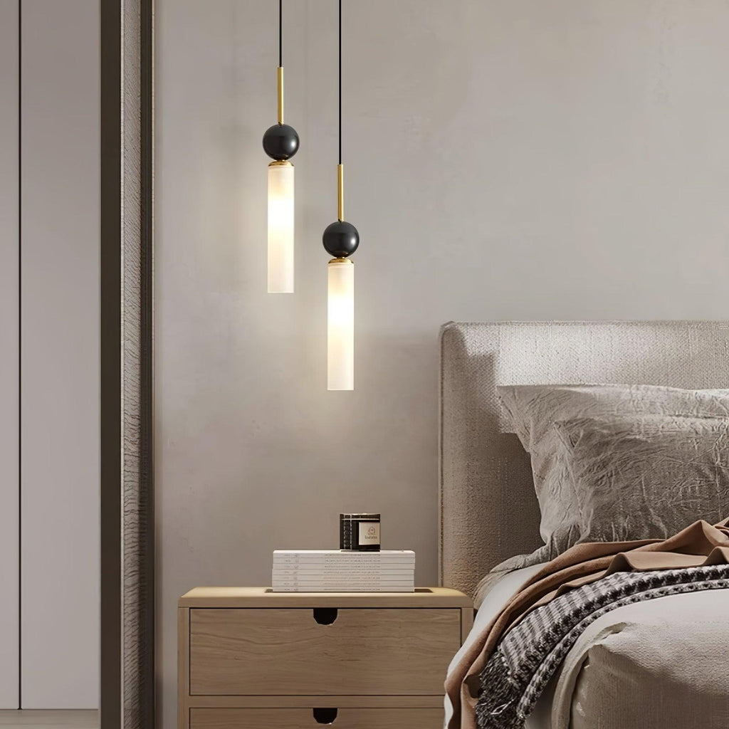 A close-up of two brass-neck Clérachic pendant lights beside an upholstered headboard and a wood nightstand holding a candle.