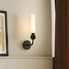A Clérachic black wall sconce lit beside a framed picture propped against a plain wall, its frosted cylindrical shade glowing softly.