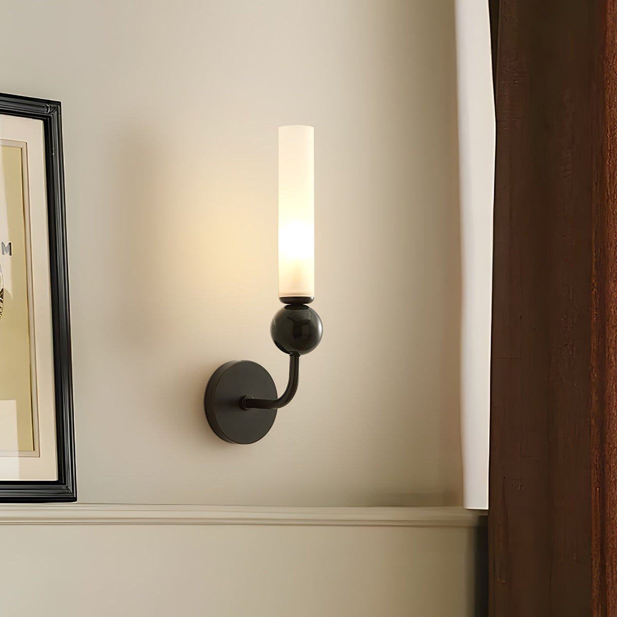 A Clérachic black wall sconce lit beside a framed picture propped against a plain wall, its frosted cylindrical shade glowing softly.