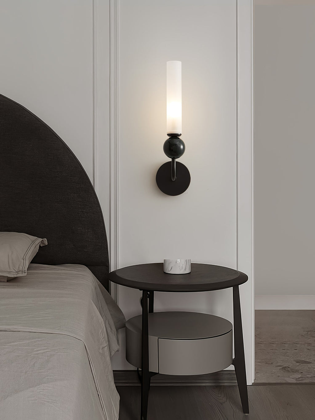 A dark round nightstand holds a cup beside a curved upholstered headboard, lit by a Clérachic black wall sconce.