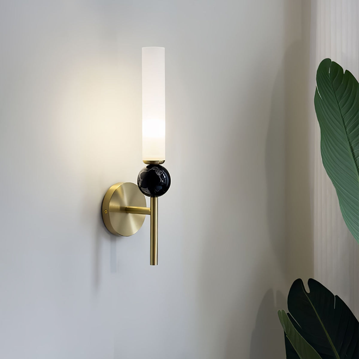 Angled shot of a Clérachic brass wall sconce with a black marble orb, a large monstera leaf visible at the edge of frame.