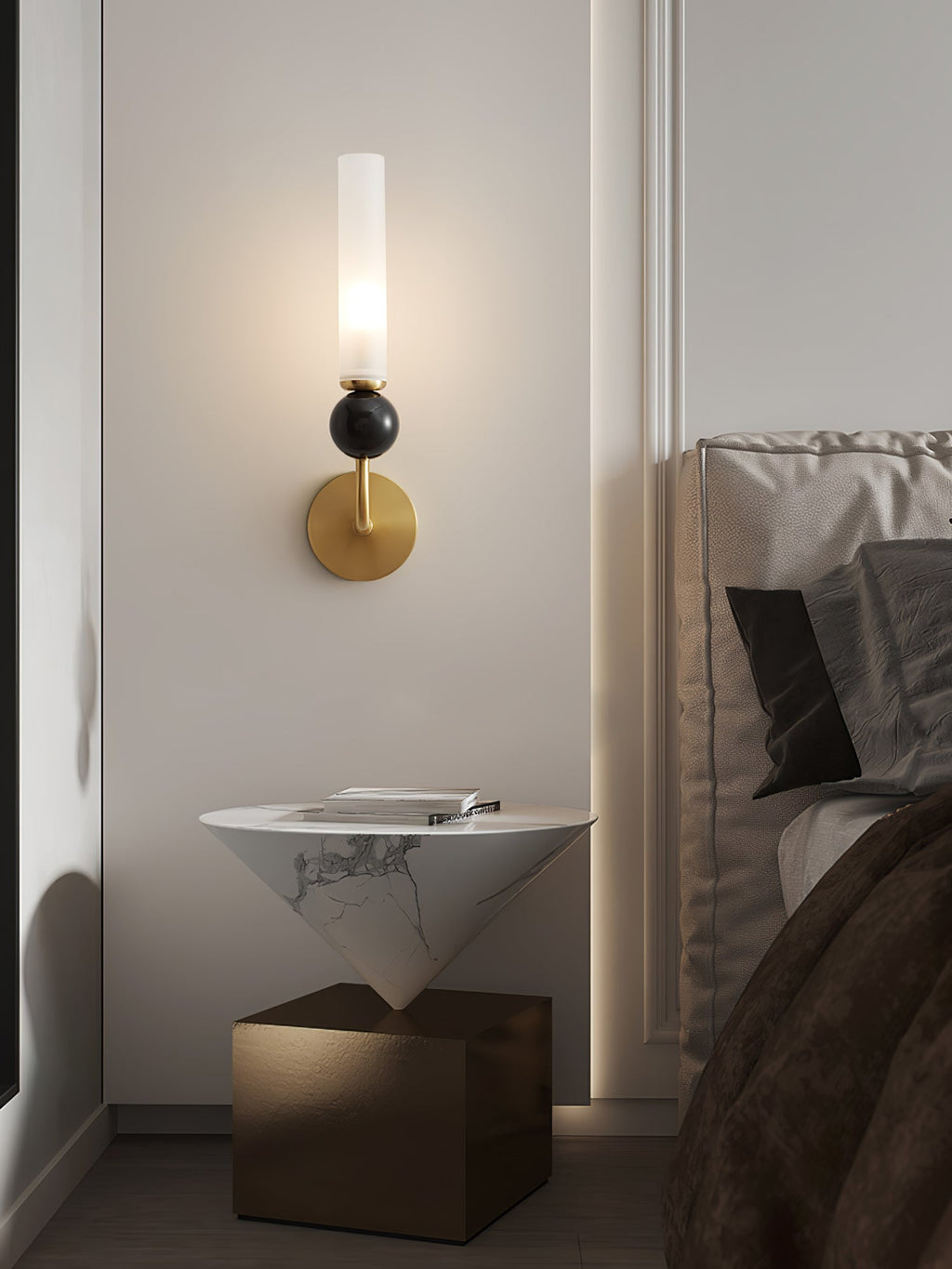 A Clérachic brass wall sconce lights a triangular marble-look side table holding a book, with a bed visible behind it.