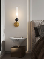 A Clérachic brass wall sconce lights a triangular marble-look side table holding a book, with a bed visible behind it.