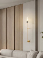 A tall Clérachic brass vertical wall light glows on a wood-panelled wall above a beige sofa in a living room.