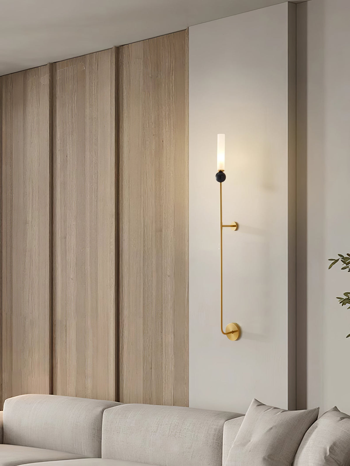 A tall Clérachic brass vertical wall light glows on a wood-panelled wall above a beige sofa in a living room.