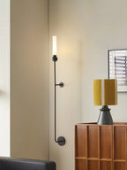 A tall Clérachic black vertical wall light stands beside a wood console table holding a mustard-shaded lamp in a living room.