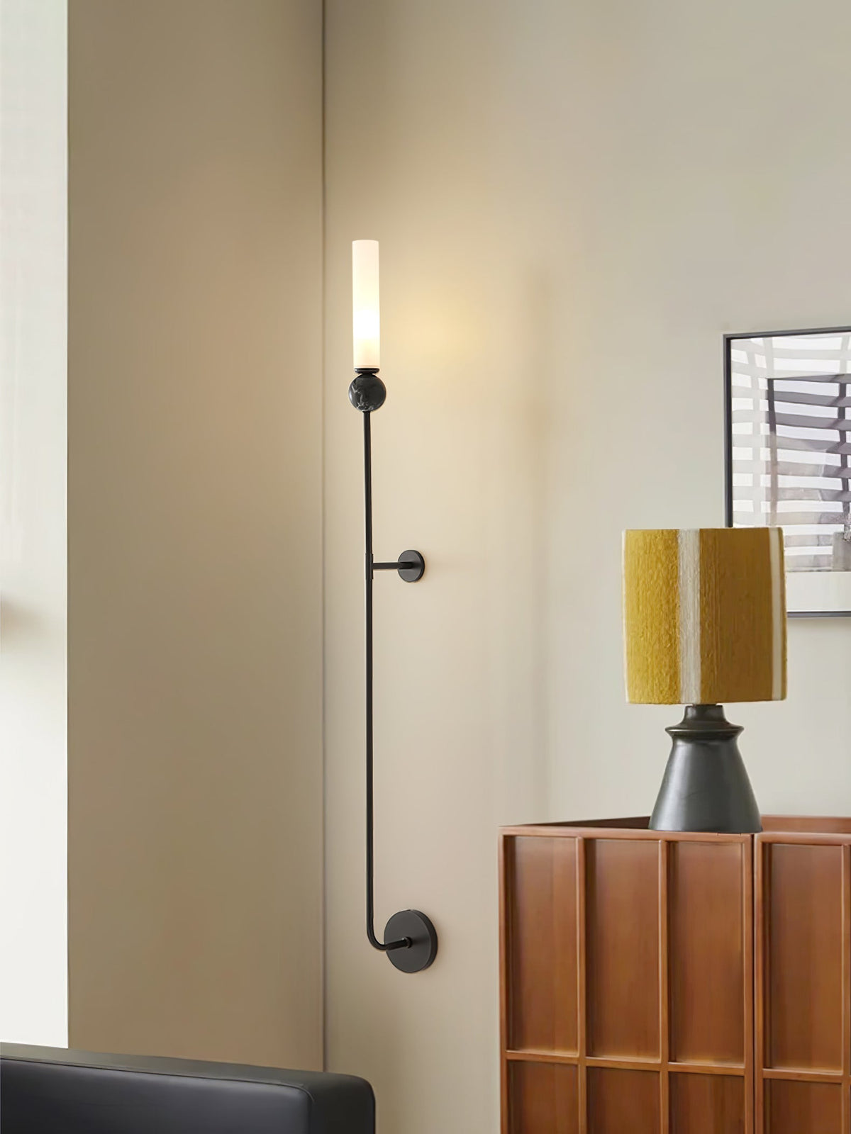 A tall Clérachic black vertical wall light stands beside a wood console table holding a mustard-shaded lamp in a living room.
