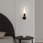 A Clérachic black wall sconce lights a small round side table beside an upholstered headboard in a cream-panelled bedroom.
