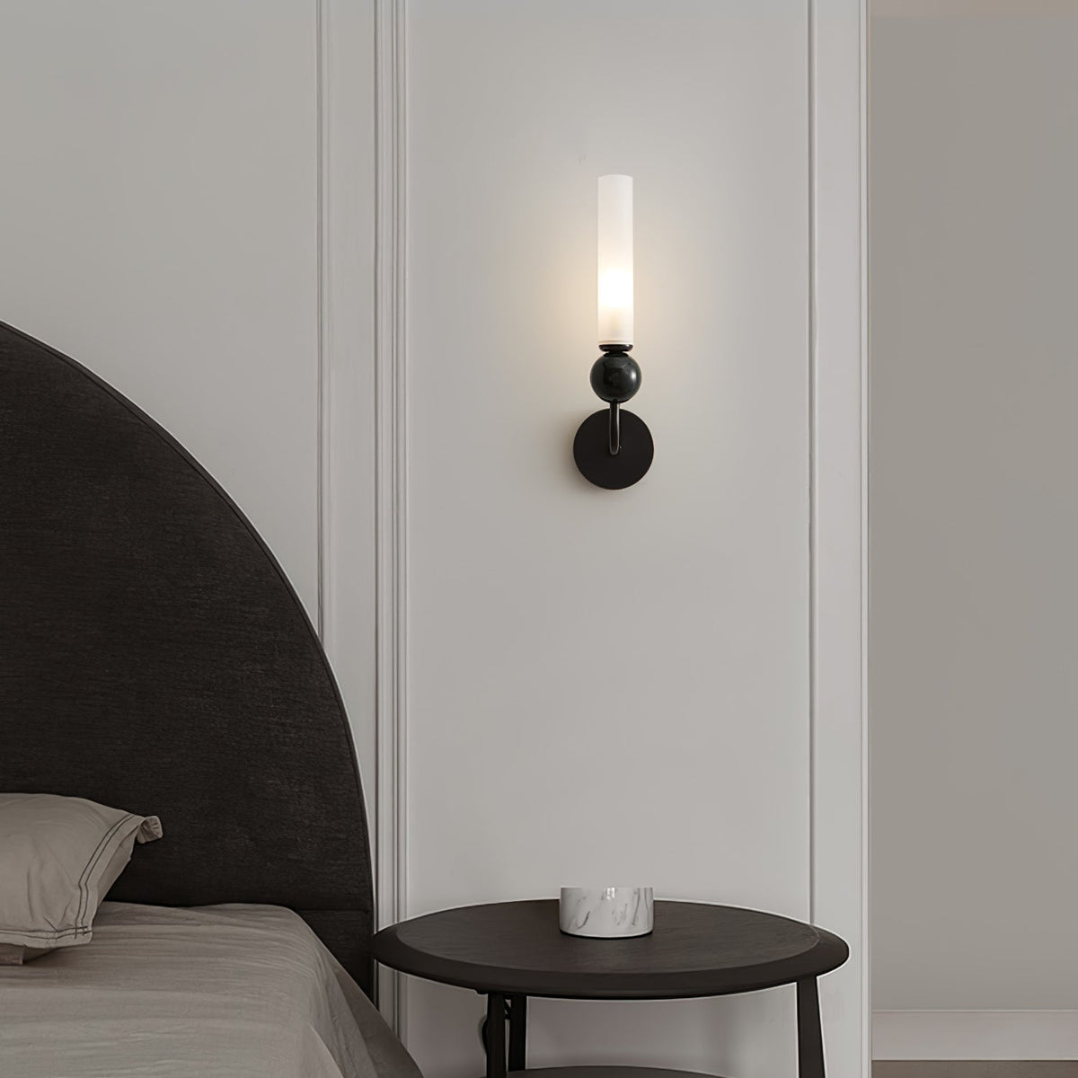 A Clérachic black wall sconce lights a small round side table beside an upholstered headboard in a cream-panelled bedroom.