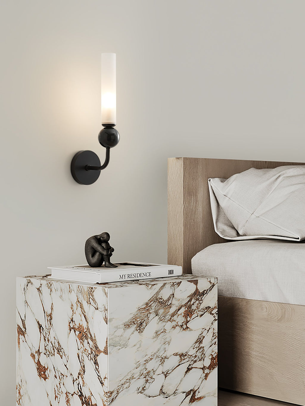 A Clérachic brass wall sconce lights a nightstand holding a small figurine and stacked books beside cream bedding.