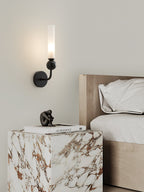 A Clérachic brass wall sconce lights a nightstand holding a small figurine and stacked books beside cream bedding.