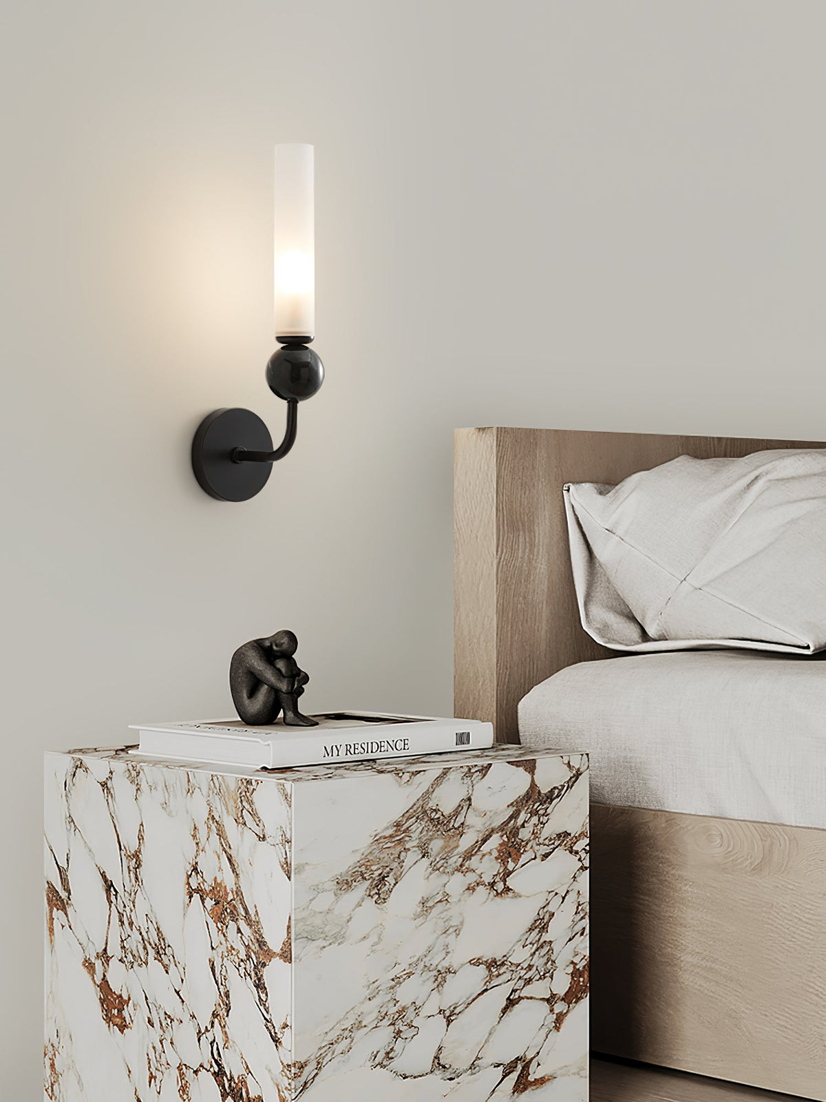 A Clérachic brass wall sconce lights a nightstand holding a small figurine and stacked books beside cream bedding.