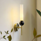 Close angle on a Clérachic brass wall sconce with a black marble orb, its warm glow set against out-of-focus leaves in the foreground.