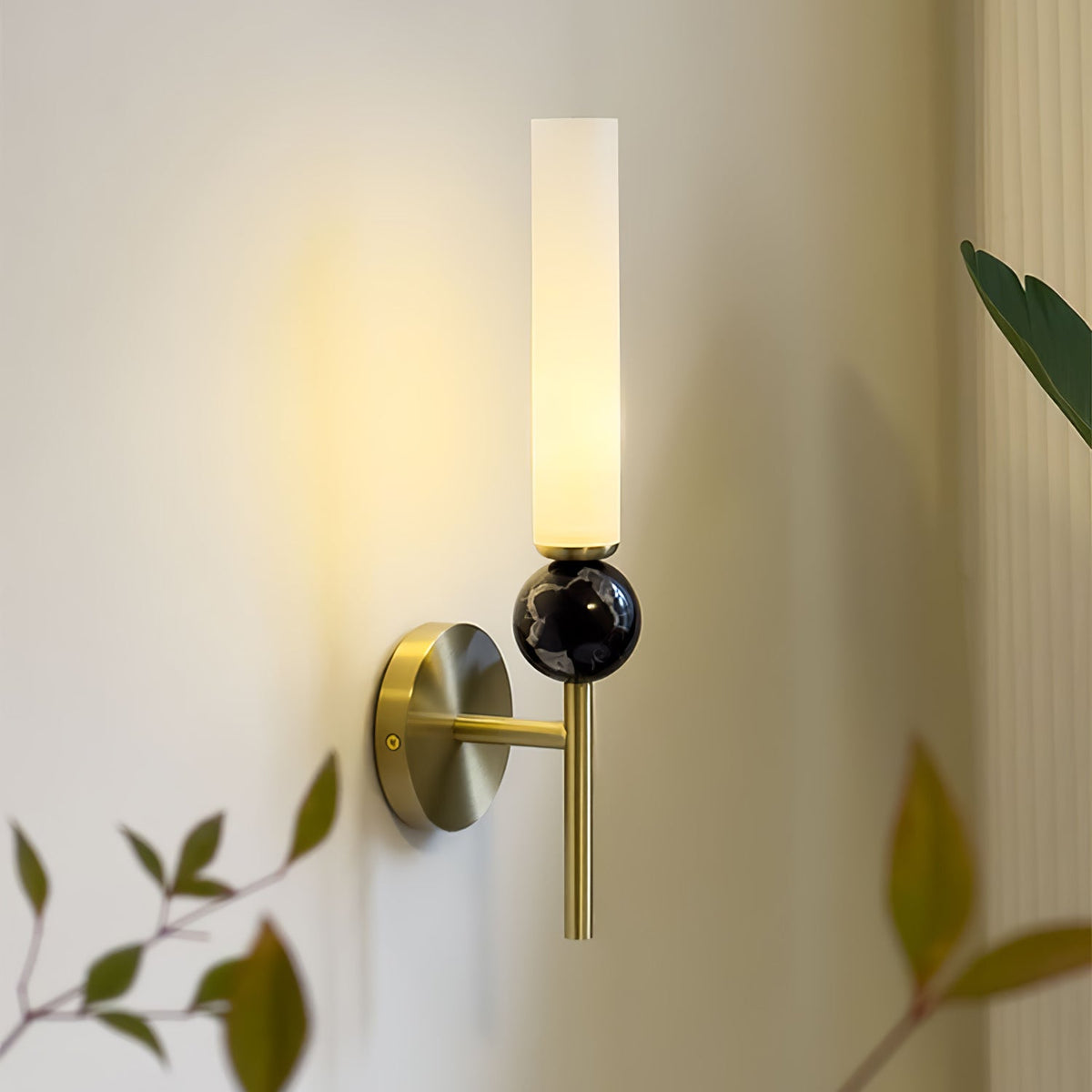Close angle on a Clérachic brass wall sconce with a black marble orb, its warm glow set against out-of-focus leaves in the foreground.