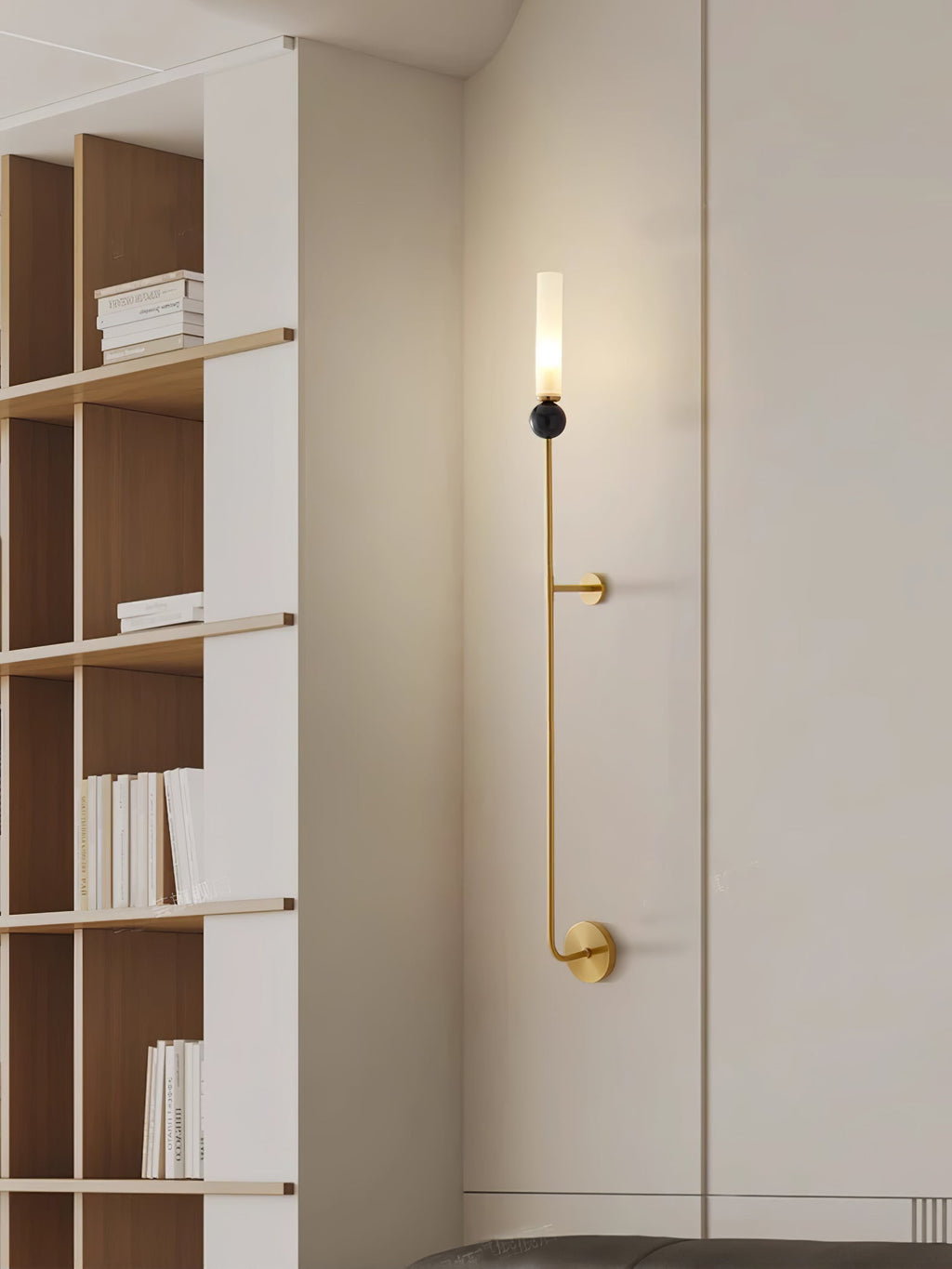 A tall Clérachic brass vertical wall light runs down a corridor wall beside built-in shelves stacked with books.
