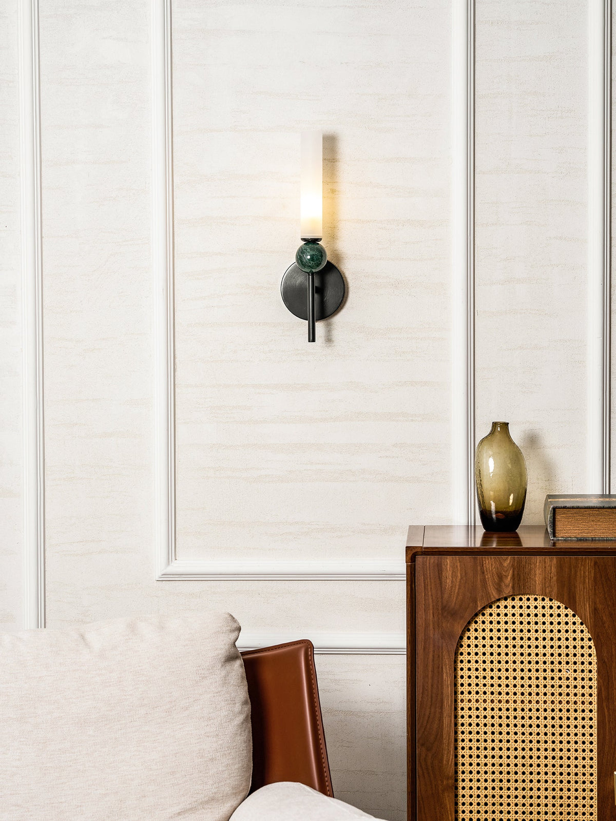 A Clérachic black wall sconce with a green marble orb glows on a panelled wall beside a rattan-fronted wooden sideboard.