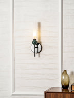 A Clérachic black wall sconce with a green marble orb glows against textured cream wallpaper near a wood table holding a vase.