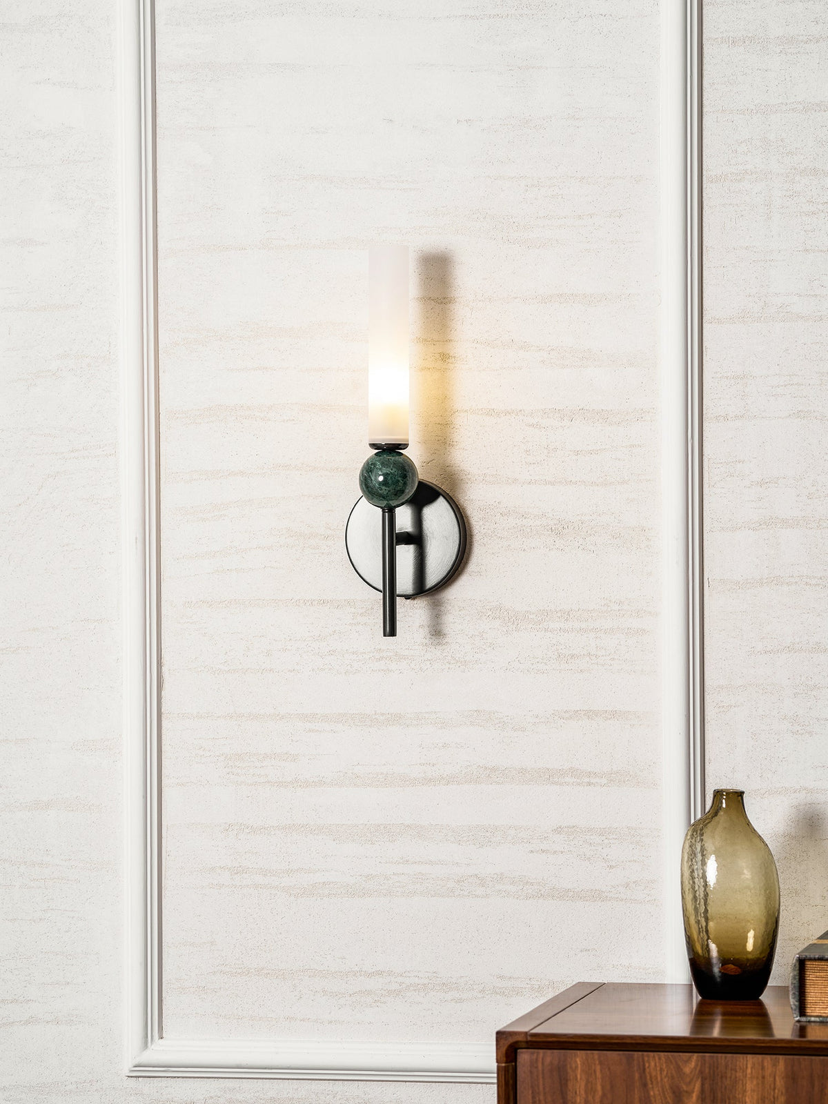 A Clérachic black wall sconce with a green marble orb glows against textured cream wallpaper near a wood table holding a vase.