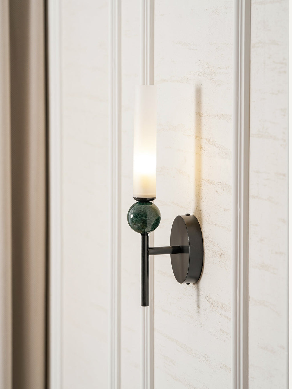The Clérachic wall light lit on a panelled wall, curtain falling to the left and its glow washing the panel behind it.