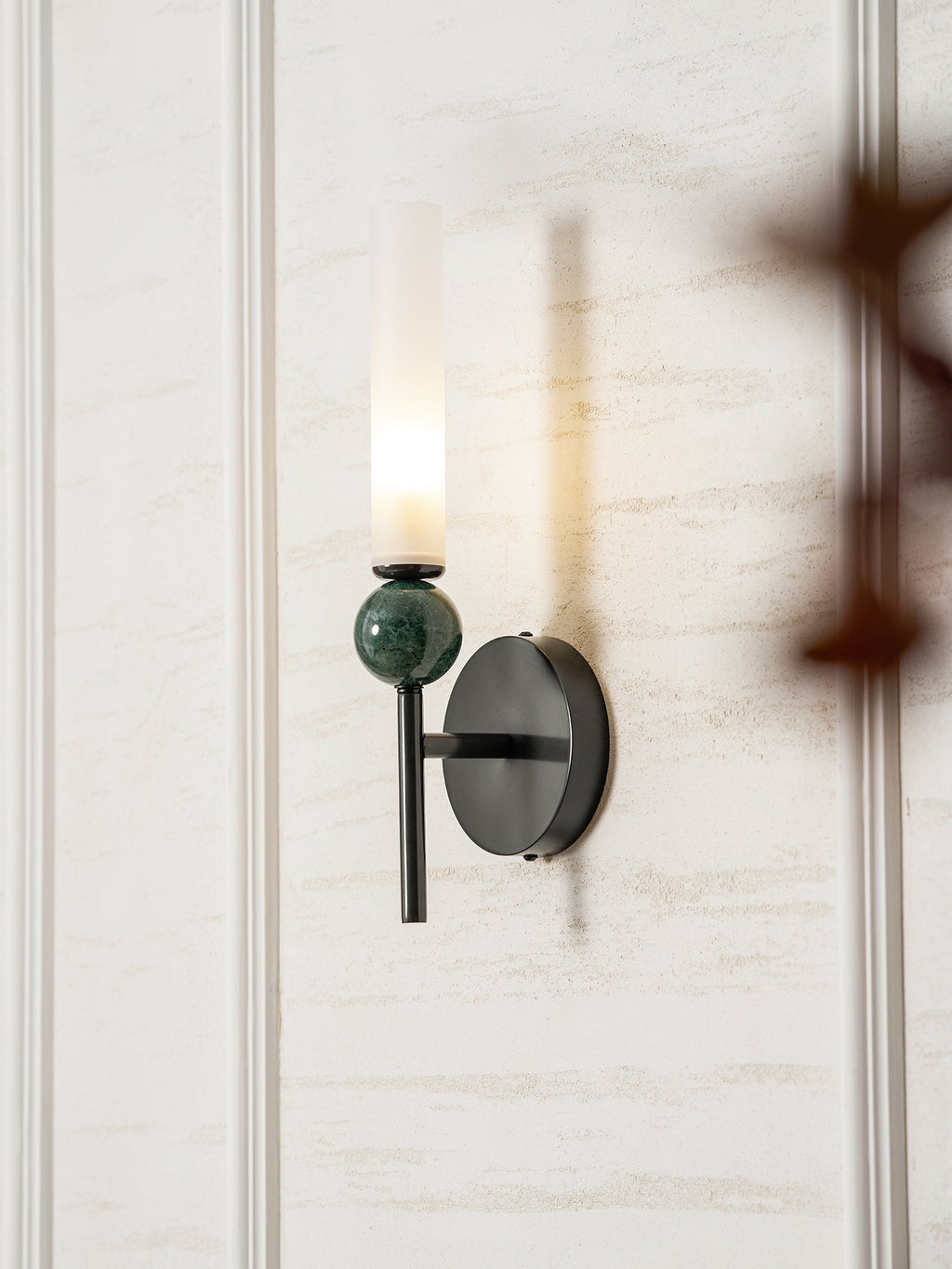 A blurred branch crosses the foreground while the Clérachic wall light glows against the panelling behind it.