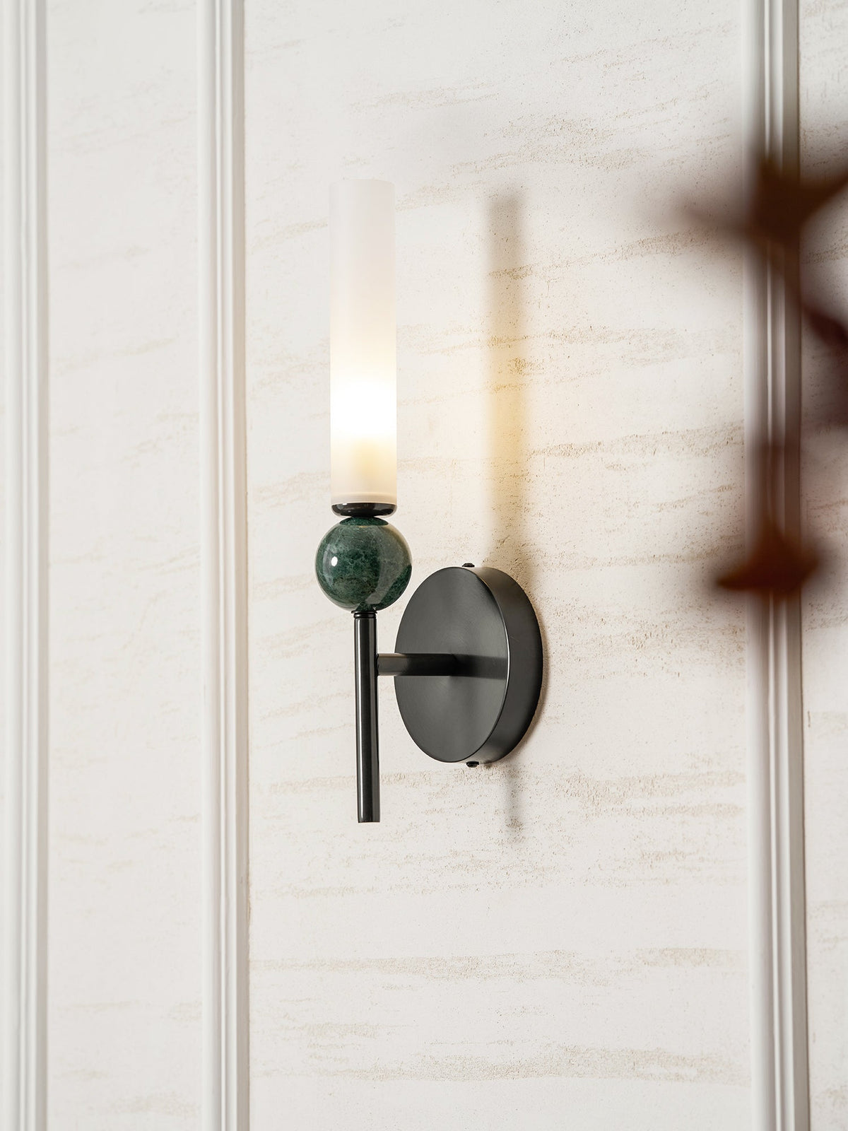 A blurred branch crosses the foreground while the Clérachic wall light glows against the panelling behind it.