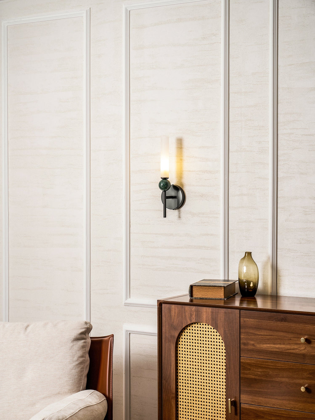 A room view with an armchair and cane cabinet, the Clérachic wall light lit small and high on the panelled wall.