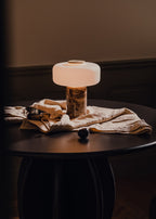 Its cord resting on a draped cloth, a Clérachic table lamp with a dark marble base is lit on a round black side table.