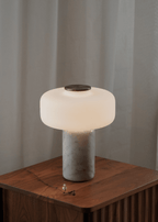 A Clérachic table lamp with a white marble base glows on a wooden nightstand in front of sheer curtains.