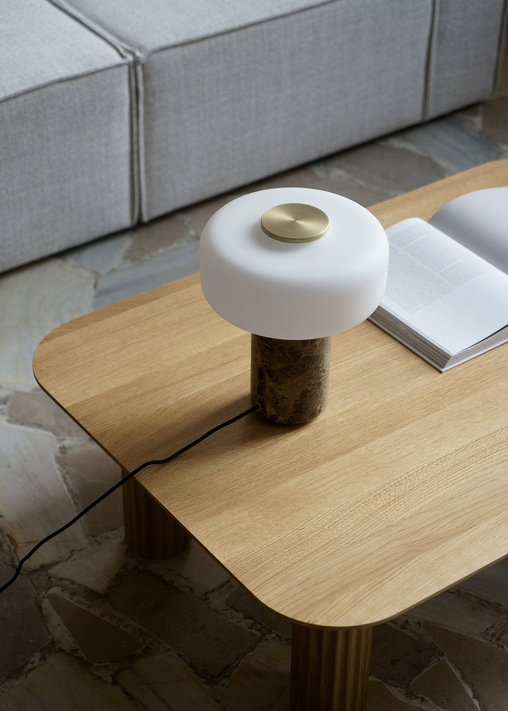 Beside an open book near a bright window, a Clérachic table lamp with a white marble base sits unlit on a wooden table.