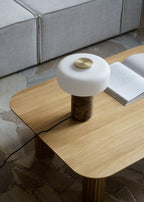 Beside an open book near a bright window, a Clérachic table lamp with a white marble base sits unlit on a wooden table.