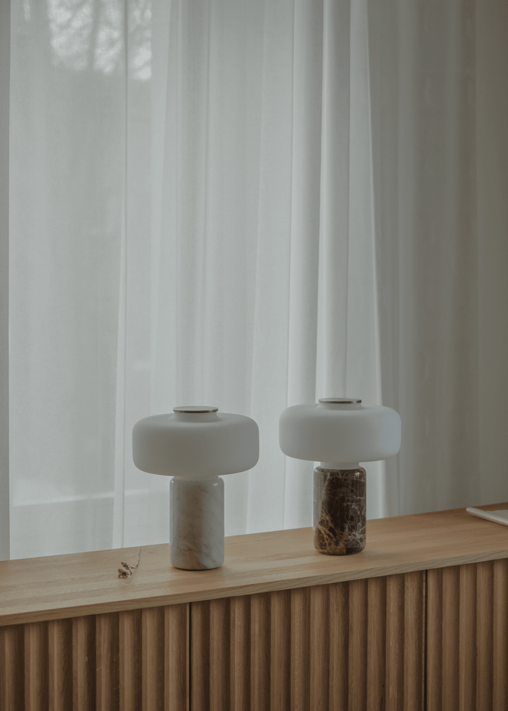 Two Clérachic table lamps, one dark marble and one white marble, glow side by side on a wooden sideboard by curtains.