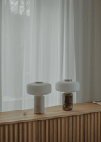Two Clérachic table lamps, one dark marble and one white marble, glow side by side on a wooden sideboard by curtains.