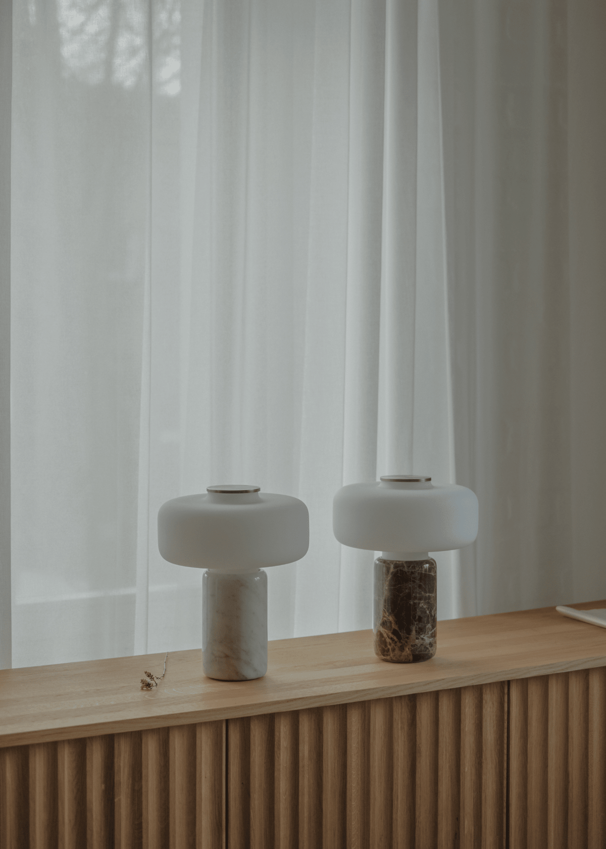 Two Clérachic table lamps, one dark marble and one white marble, glow side by side on a wooden sideboard by curtains.
