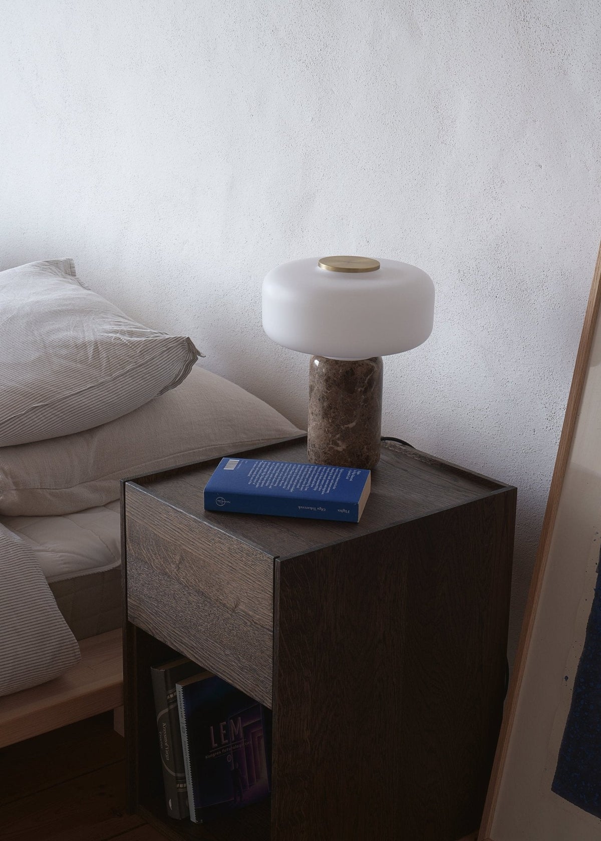 A book stacked below it, a Clérachic table lamp with a marbled base is lit on a dark wooden nightstand beside a bed.