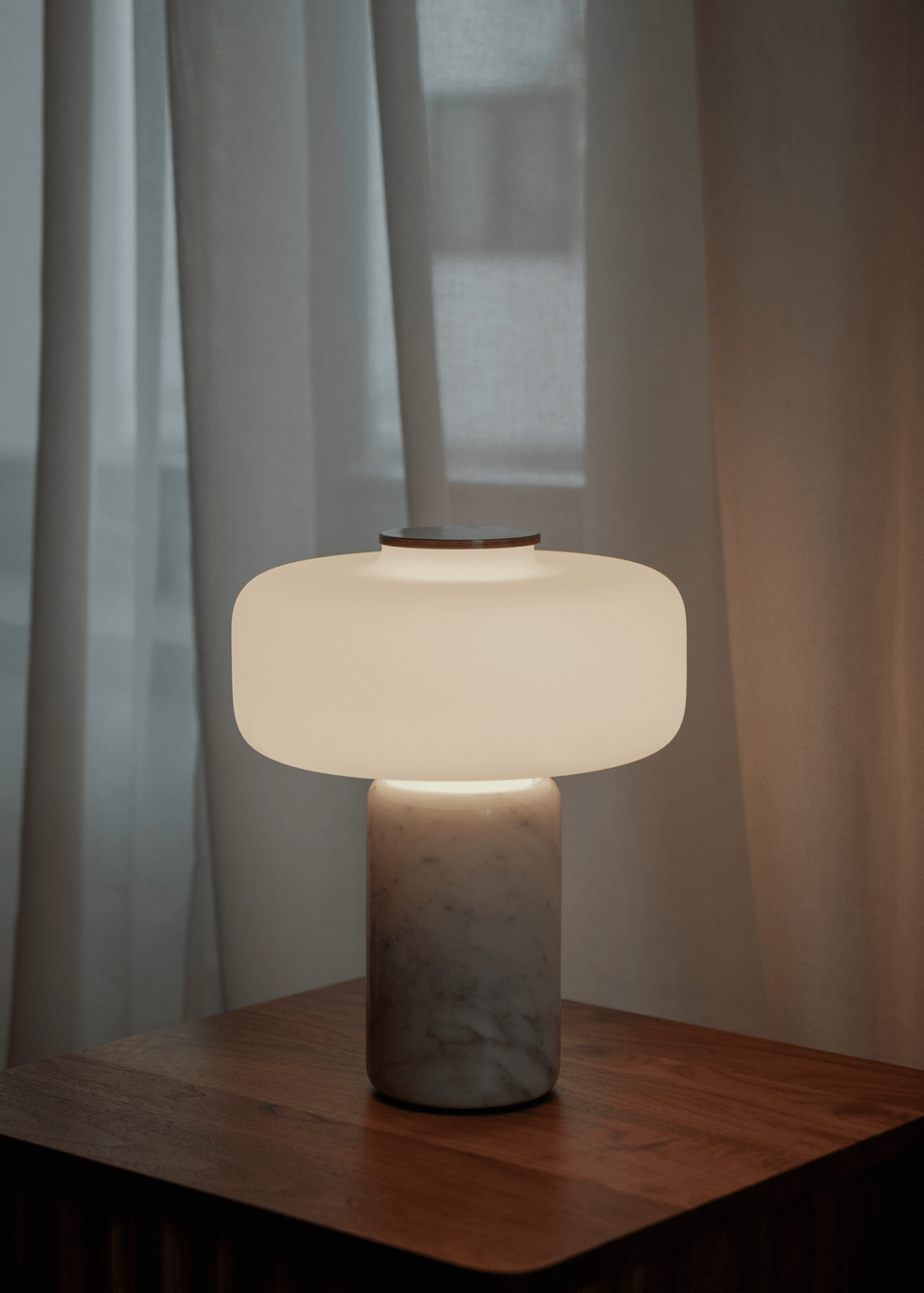 A Clérachic table lamp with a white marble base glows on a wooden nightstand against sheer curtains at dusk.