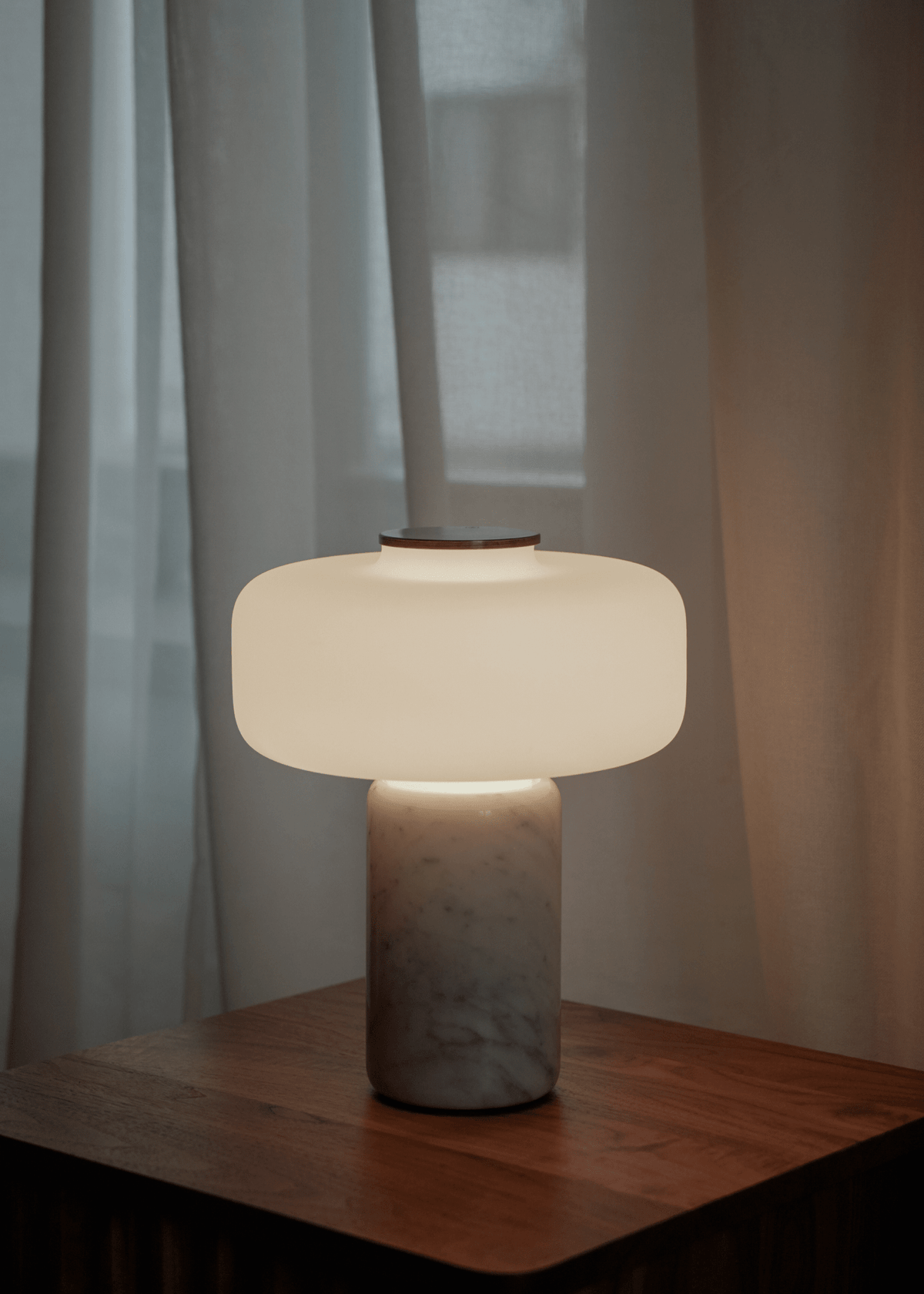 A Clérachic table lamp with a white marble base glows on a wooden nightstand against sheer curtains at dusk.