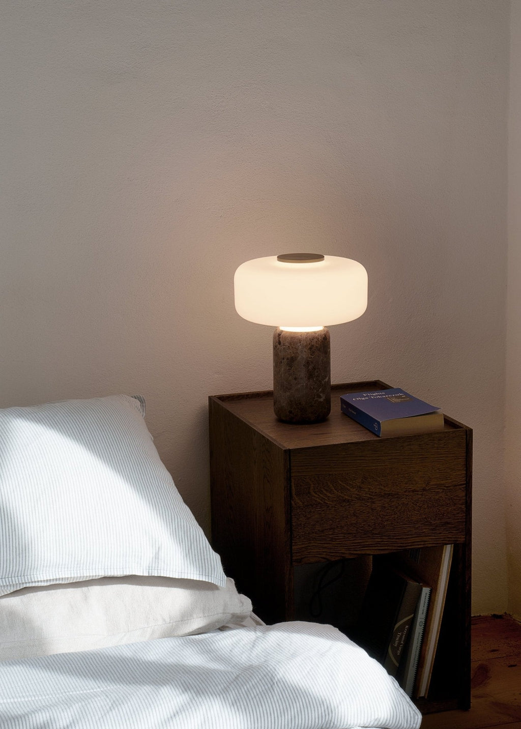 Beside a white-linened bed, a Clérachic table lamp with a dark marble base glows warmly on the wooden nightstand.