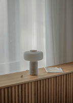 On a wooden windowsill shelf in front of sheer curtains, a Clérachic table lamp with a pale marble base glows faintly.