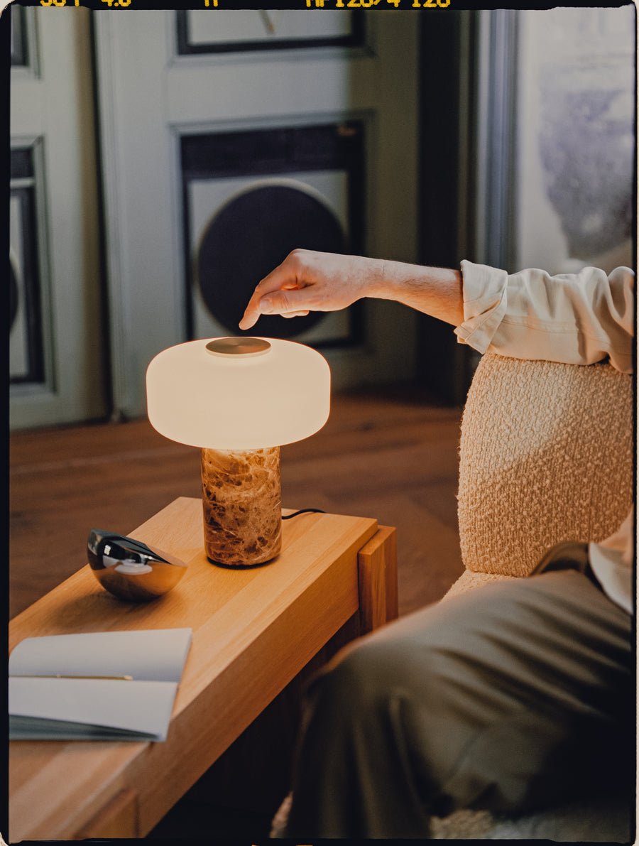 A hand reaches toward the touch switch of a Clérachic table lamp with a dark marble base, lit on a nightstand at night.