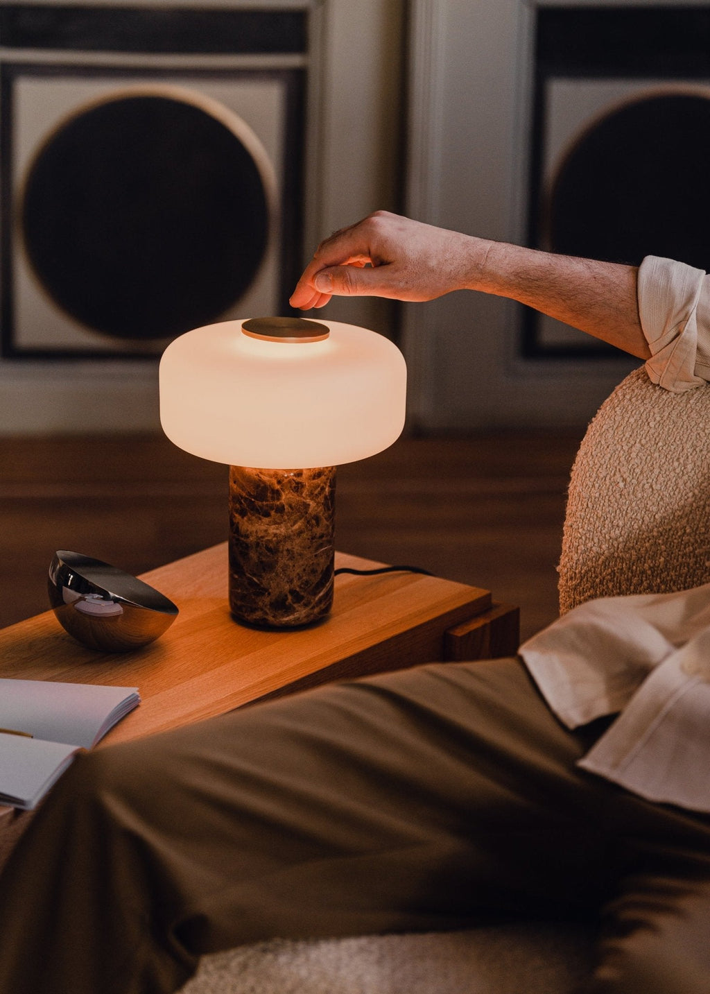 A hand reaches to touch the brass switch on a Clérachic table lamp with a dark marble base, lit beside a bed at night.