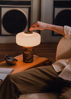 A hand reaches to touch the brass switch on a Clérachic table lamp with a dark marble base, lit beside a bed at night.