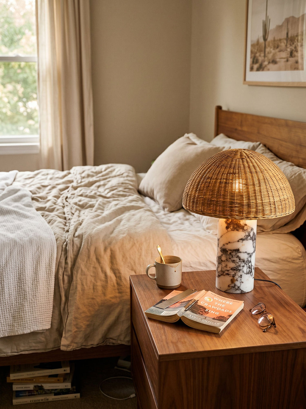 The rattan dome lamp from Clérachic glowing on a bedside table beside white linen bedding and reading glasses.