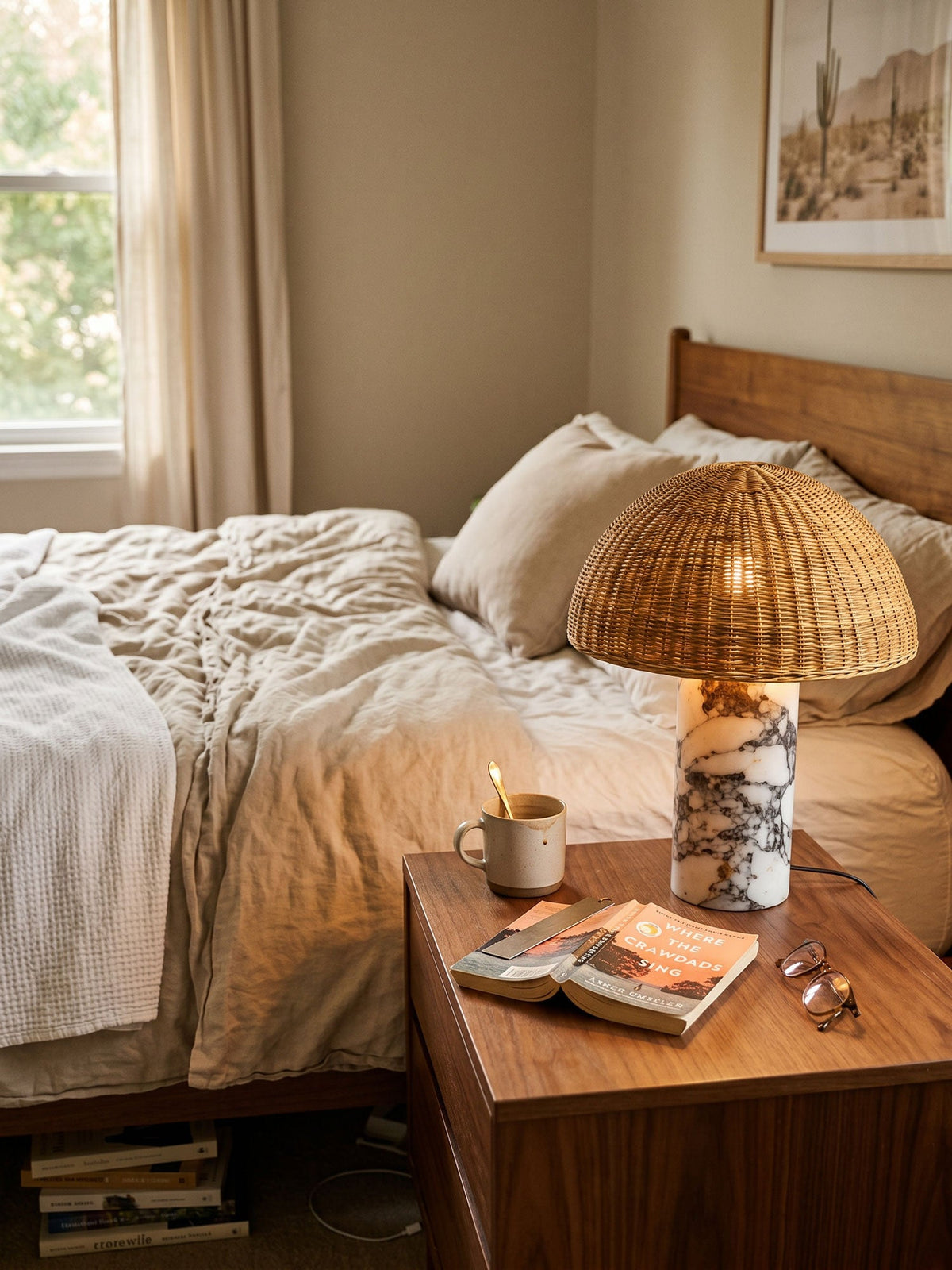 The rattan dome lamp from Clérachic glowing on a bedside table beside white linen bedding and reading glasses.