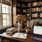Clérachic's rattan table lamp lit on a wooden desk beside an open notebook, a laptop and a stack of books.