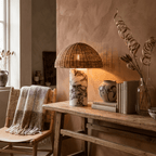 Clérachic's woven rattan table lamp glowing on a wooden console with dried branches and a small ceramic vase.