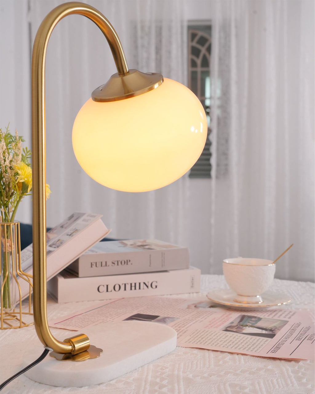 The Clérachic table lamp's white shade glows on a gold arm beside a stacked magazine, teacup and pink accessories on a wooden table.