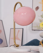 An unlit pink-shaded Clérachic table lamp on a gold arm stands against a dark abstract artwork beside a blue cushion.