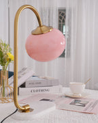 The Clérachic table lamp's pink shade sits unlit on a gold arm beside a small vase of white flowers and a stack of magazines.