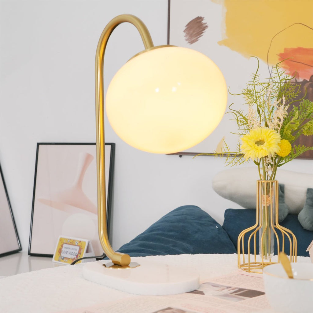 A lit Clérachic table lamp with brass frame and white shade sits on a bed, framed art and a wire vase of dried flowers beside a navy cushion.